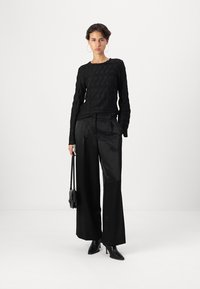 Maglietta nera a maniche lunghe con motivo testurizzato, abbinata a pantaloni ampi a vita alta. La modella tiene una piccola borsa nera e indossa tacchi neri.