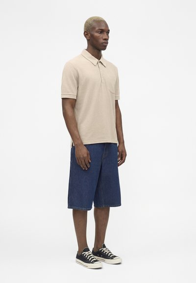 Άνδρας στέκεται φορώντας μπέζ πόλο με κοντά μανίκια, μπλε τζιν σορτς μέχρι το γόνατο και μαύρα χαμηλά αθλητικά παπούτσια με λευκά κορδόνια.