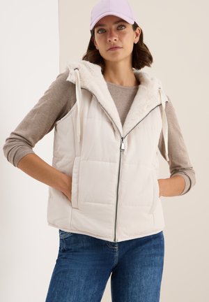 Gilet blanc moelleux avec capuche en fourrure, fermeture éclair et poches avant, associé à une chemise beige à manches longues et un jean bleu.