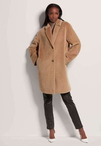 Beige oversized wollen jas met een opstaand kraag en twee knopen, gestyled over een lichte coltrui. Gecombineerd met zwarte textuur broek en witte hakken.