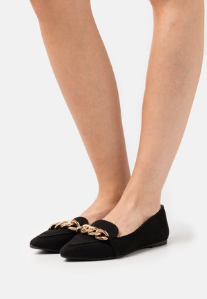 Mocassins en suede noir avec un bout pointu et une large sangle ornée de chaînes dorées à l'avant. Semelle plate, design minimaliste.