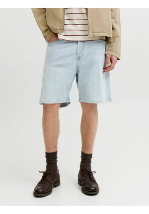 Mężczyzna w jasnoniebieskich dżinsowych szortach, beżowej kurtce, pasiastym t-shircie, ciemnobrązowych skarpetach i ciemnobrązowych sznurowanych butach, stojący z rękami w kieszeniach.