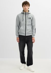 Sudadera gris con cremallera, capucha ajustable, bolsillos frontales y detalle del logo en negro. Combinada con jeans oscuros y zapatillas blancas.