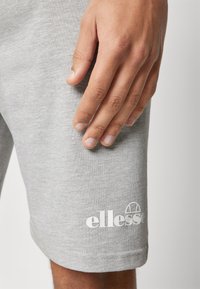 Grijze katoenen sportshorts met een gladde textuur, voorzien van een wit "ellesse" logo nabij de zoom en een opgerolde zoomdetail.