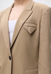 Close-up af beige blazer med unikt trekantet foldet lomme detalje over en hvid top.