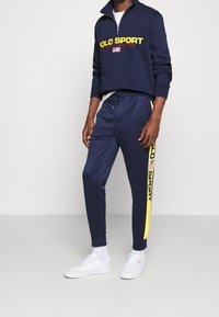 Dunkelblauer Sweatshirt mit gelbem Text und Logo, Reißverschluss am Hals; dunkelblaue Jogginghosen mit gelben Seitenstreifen und Logo; weiße Sneaker.