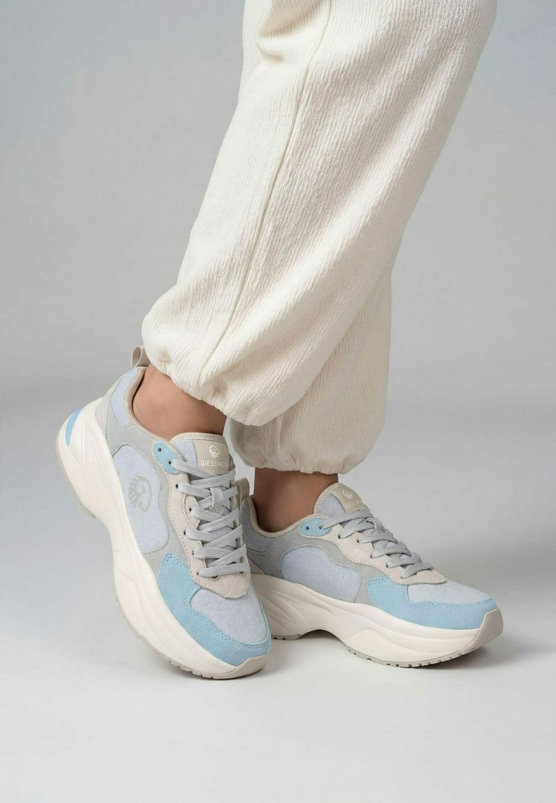 Voeten met witte en lichtblauwe chunky sneakers, gecombineerd met crème textuurbroeken met elastische boorden, staand op een effen lichte ondergrond.