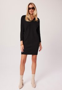 Czarna dzianinowa sukienka z prążkowanego materiału, z dekoltem w serek, długimi rękawami i długością do połowy uda. Noszona z beżowymi botkami na obcasie i brązowymi okularami przeciwsłonecznymi.