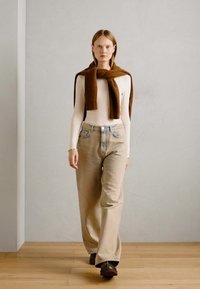 Beige wide-leg jeans, white ribbed long-sleeve top, and a brown knit scarf tied around the neck. Model walking on wooden floor against gray backdrop.