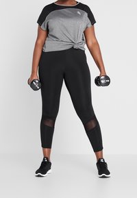 Sportsoutfit med en grå og sort kortærmet top, sorte leggings med mesh-detaljer og sorte sneakers, som holder håndvægte.