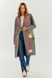 Kolorowy płaszcz w kratę houndstooth z różowymi, pomarańczowymi i żółtymi paskami; hafty kwiatowe; otwarty przód; średniej długości; połączony z niebieskimi dżinsami i trampkami.