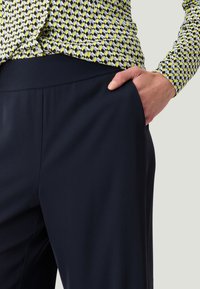 Marineblauwe broek met een gladde textuur, voorzien van een brede tailleband en zijzakken. Een gestreept bovenstuk in geel, zwart en wit is gedeeltelijk zichtbaar.