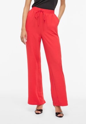 Γυναίκα με φωτεινά κόκκινα φαρδιά παντελόνια με κορδόνι στη μέση και μαύρες ανοιχτές γόβες, στέκεται με το ένα χέρι στην τσέπη.