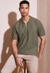 Olivově zelené polo triko s krátkým rukávem, s límcem na zip, texturovaná látka, přiléhavý střih, nošené s béžovými kalhotami, sedící na dřevěné stoličce.