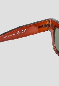 Lunettes de soleil transparentes marron avec des verres vert foncé. Présente un liseré noir et une charnière en métal. Le texte sur la branche inclut « FABRIQUÉ EN CHINE » et le marquage CE.