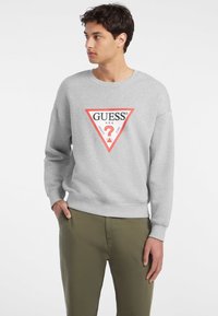 Grå hettegenser laget av mykt stoff, med et rødt trekantet logo som har "GUESS" og et spørsmålstegn inni. Lange ermer og ribbede mansjetter.