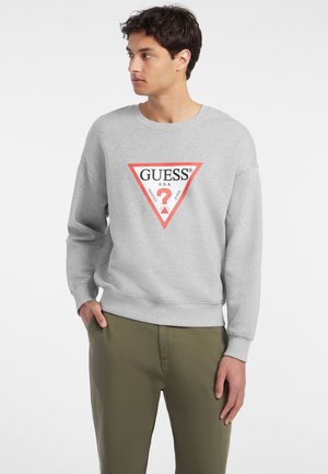 Felpa grigia in tessuto morbido, con un logo triangolare rosso che presenta la scritta "GUESS" e un punto interrogativo all'interno. Maniche lunghe e polsini a costine.