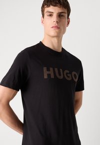 Melnā T-kreklā ar īsām piedurknēm, pārsteidz ar lielu brūnu "HUGO" logotipu priekšpusē. Izgatavots no gludas auduma, tam ir apaļš kakls.
