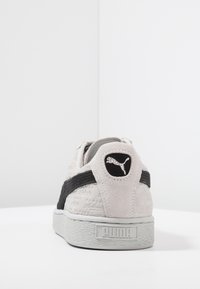 Vaaleanharmaat mokkasiinit, joissa on teksturoidut yksityiskohdat, musta korosteväri ja pyöreä logo-merkki kantapäällä. Kumipohja, jossa on "PUMA"-brändäys.