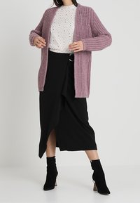 Žena v lila objemném pleteném kardiganu, bílé vzorované blouse, černé zavinovací sukni a černých kotníkových botách stojí na jednobarevném pozadí.