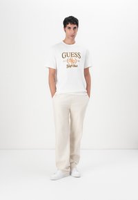 Camiseta de algodón blanca con gráfica dorada y marrón de "GUESS Golf Club". Usada con pantalones beige claros y zapatillas blancas. Diseño simple, corte relajado.