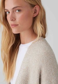 Cardigan beige chiaro con trama, dalla superficie morbida e pelosa e fili metallici, indossato sopra una maglietta bianca. Taglio dritto e design aperto.