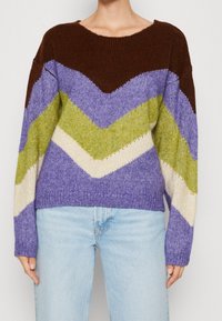 Knit sweater with chevron stripes in brown, purple, green, and cream worn with light blue jeans, shown from neck to hips.