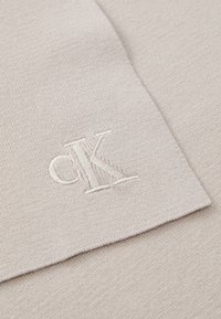 Beez žaketkangas, millel on esiletungiv valge tikkitud "ck" logo. Tekstuur on sile ja delikaatsete soontega.