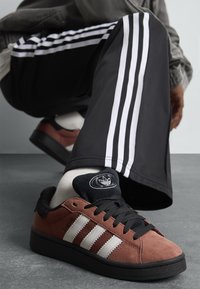 Brown suede sneakers with black accents and white three-stripe design, featuring black laces and a textured sole. White socks and black pants visible.