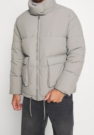 Homme portant une doudoune gris clair avec fermeture éclair sur le devant, grandes poches à rabat et cordons ajustables à l'ourlet, assortie à un jean foncé.