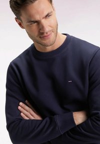 Homme avec une légère barbe portant un sweat-shirt bleu marine, les bras croisés, regardant légèrement vers le bas sur un fond clair.