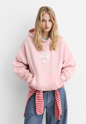 Giovane donna con lunghi capelli biondi che indossa un hoodie rosa con il testo "IN A DREAM", una maglietta a righe legata in vita e jeans blu, in piedi contro un muro bianco.