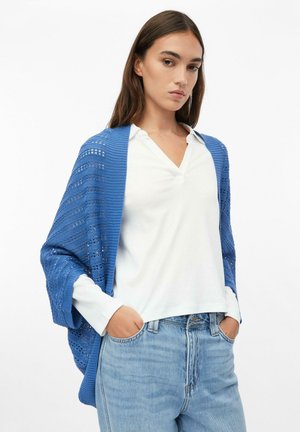 Frau trägt einen blauen gestrickten Schal über einer weißen Bluse und hellblauen Jeans, die Hände in den Taschen, schaut mit neutralem Gesichtsausdruck nach vorn.