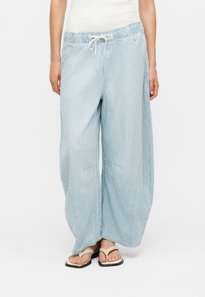 Γυναίκα που φοράει ανοιχτομπλε και λευκή κάθετη ριγέ παντελόνια με φαρδιά πόδια, ρυθμιζόμενο ύψος στη μέση και λευκά σαγιονάρες.
