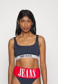 Námornícky modrý crop top s bielym p�ásom „TOMMY JEANS“; v kombinácii s vysokými červenými nohavičkami s veľkým bielym nápisom „JEANS“. Hladká textúra.