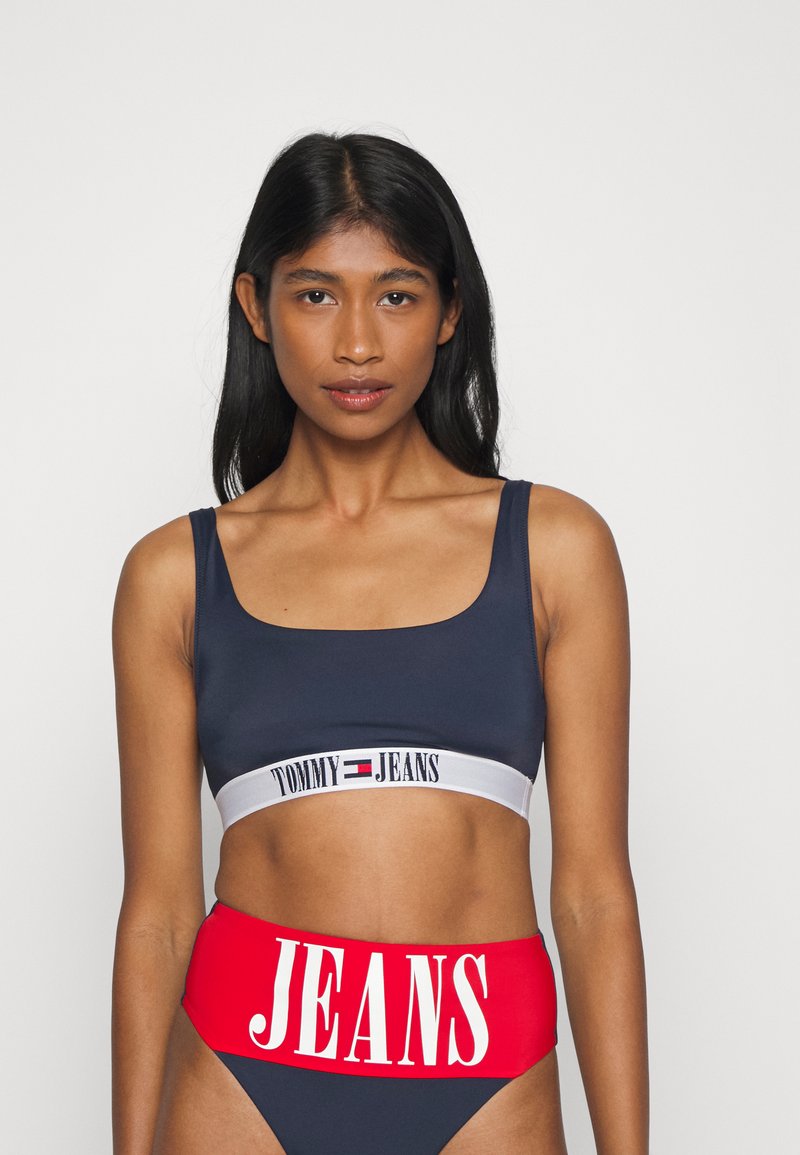 Námornícky modrý crop top s bielym pásom „TOMMY JEANS“; v kombinácii s vysokými červenými nohavičkami s veľkým bielym nápisom „JEANS“. Hladká textúra.
