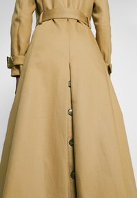 Trench-coat beige avec une ceinture à la taille, des boutons dans le dos et des lanières aux poignets des manches longues.