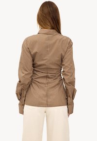 Brown striped button-down shirt with a cinched back design, long sleeves, and a classic collar, paired with light-colored pants.