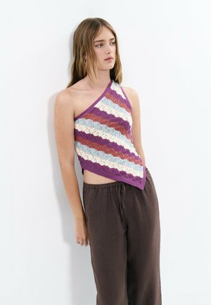 Jeune femme appuyée contre un mur blanc portant un top en maille multicolore à une épaule et un pantalon ample marron foncé avec un cordon.