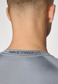 Camiseta atlética gris con una textura suave y el logo "NIKE PRO" en la parte posterior del cuello. Presenta un diseño ajustado y mangas raglán para mayor movilidad.