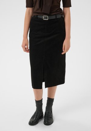 Femme portant une jupe midi en velours côtelé noire avec une fente à l'avant, une ceinture noire, des chaussettes grises et des mocassins en cuir noirs sur un fond uni.