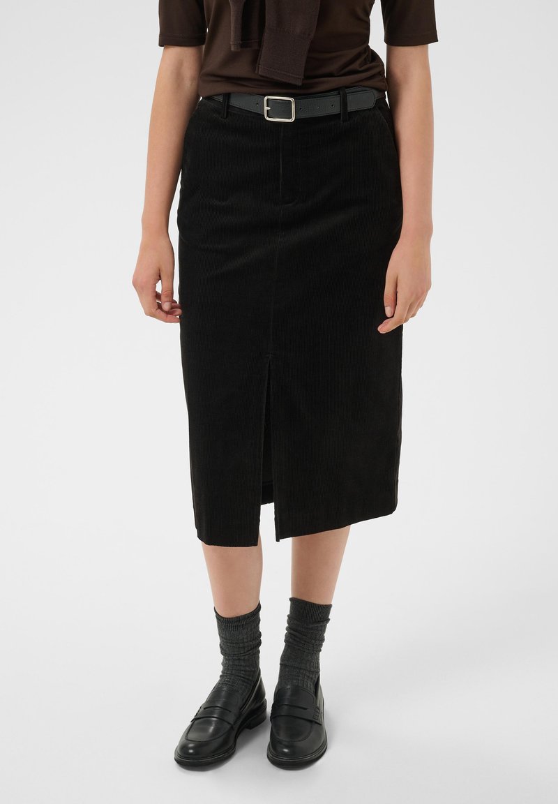 Femme portant une jupe midi en velours côtelé noire avec une fente à l'avant, une ceinture noire, des chaussettes grises et des mocassins en cuir noirs sur un fond uni.