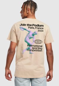 Beige bavlněné tričko s pixelovaným grafickým motivem postavy házející disky. Text obsahuje "Připojte se k Podium, Paříž, Francie 2024."
