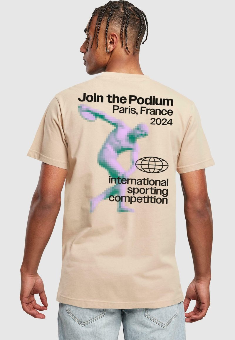 Beige bavlněné tričko s pixelovaným grafickým motivem postavy házející disky. Text obsahuje "Připojte se k Podium, Paříž, Francie 2024."