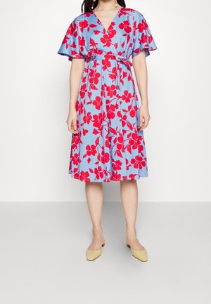 Vrouw draagt een lichtblauwe jurk tot op de knie met grote rode bloemenpatronen, korte wijd uitlopende mouwen, V-hals, tailleband en beige puntige platte schoenen.