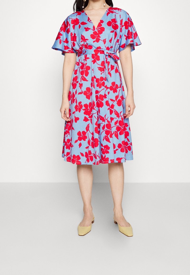 Kobieta ubrana w błękitną sukienkę do kolan z dużym czerwonym kwiatowym wzorem, krótkimi falującymi rękawami, dekoltem w serek, wiązanym w talii paskiem oraz beżowymi, spiczastymi, płaskimi butami.