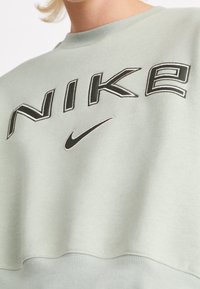 Grön sweatshirt med ribbad halsringning och muddar, som har stor broderad "NIKE"-text och logotyp i kontrasterande färger. Mjuk bomullsmaterial.