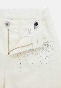 Pantaloni in denim bianco con chiusura a zip e bottone. Presentano una tasca frontale con decorazioni in strass argento vicino alla vita.