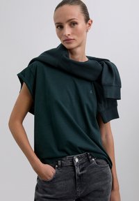 Dark green short-sleeve t-shirt with a relaxed fit, features a crew neck and has a matching sweater draped over the shoulders. Accompanied by gray jeans.