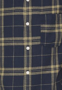 Camisa de franela a cuadros en azul marino y verde aceituna con frontales de botón, que cuenta con un bolsillo en el lado izquierdo del pecho y botones blancos. Tejido texturizado.